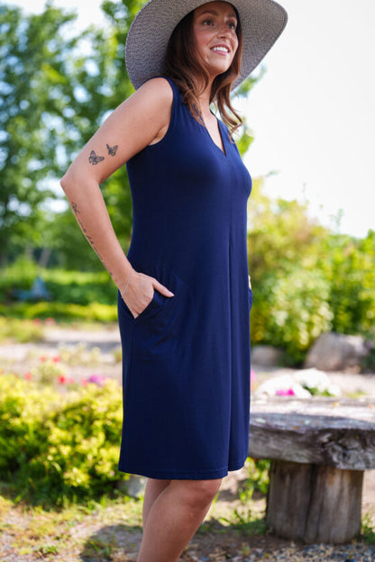 3/4 side view of a woman wearing the Samba Dress by Rien ne se Perd in Navy, a sleeveless short dress with a V-neck, centre pleat, and side pockets. She is standing outside.