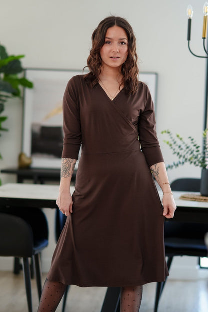 A woman wearing the St-Laurent Dress by Rien ne se Perd in Chocolate, a midi-length wrap style dress with a 3/4 sleeves and subtle shoulder pleats. She is standing in a dining room in front of a table. 