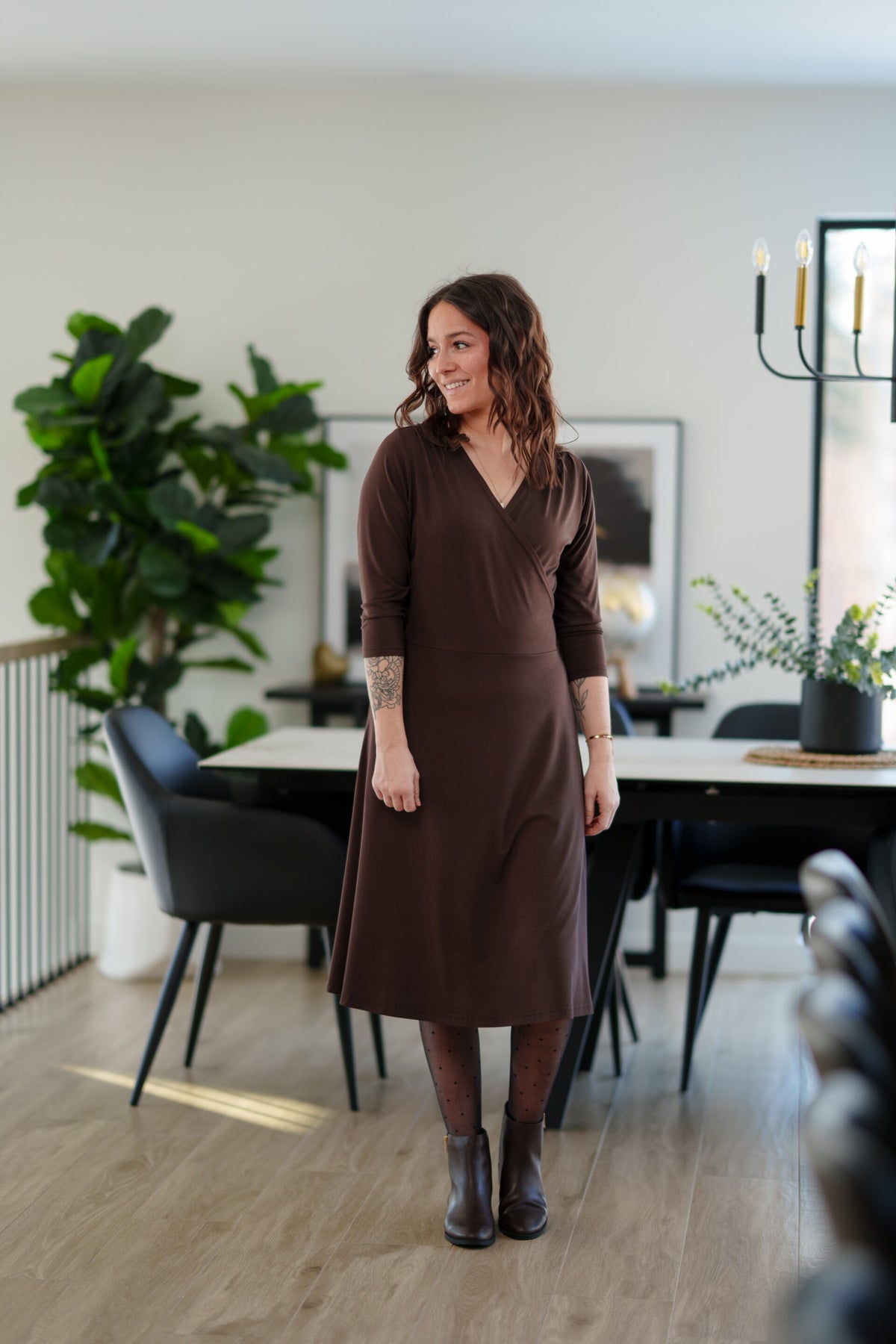A woman wearing the St-Laurent Dress by Rien ne se Perd in Chocolate, a midi-length wrap style dress with a 3/4 sleeves and subtle shoulder pleats. She is standing in a dining room in front of a table. 