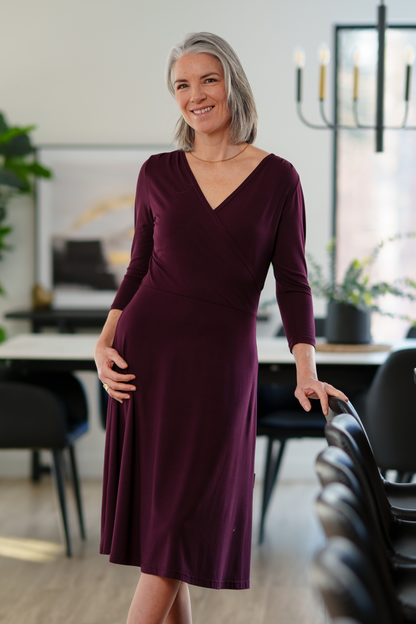 A woman wearing the St-Laurent Dress by Rien ne se Perd in Porto, a midi-length wrap style dress with a 3/4 sleeves and subtle shoulder pleats. She is standing in a dining room in front of a table. 