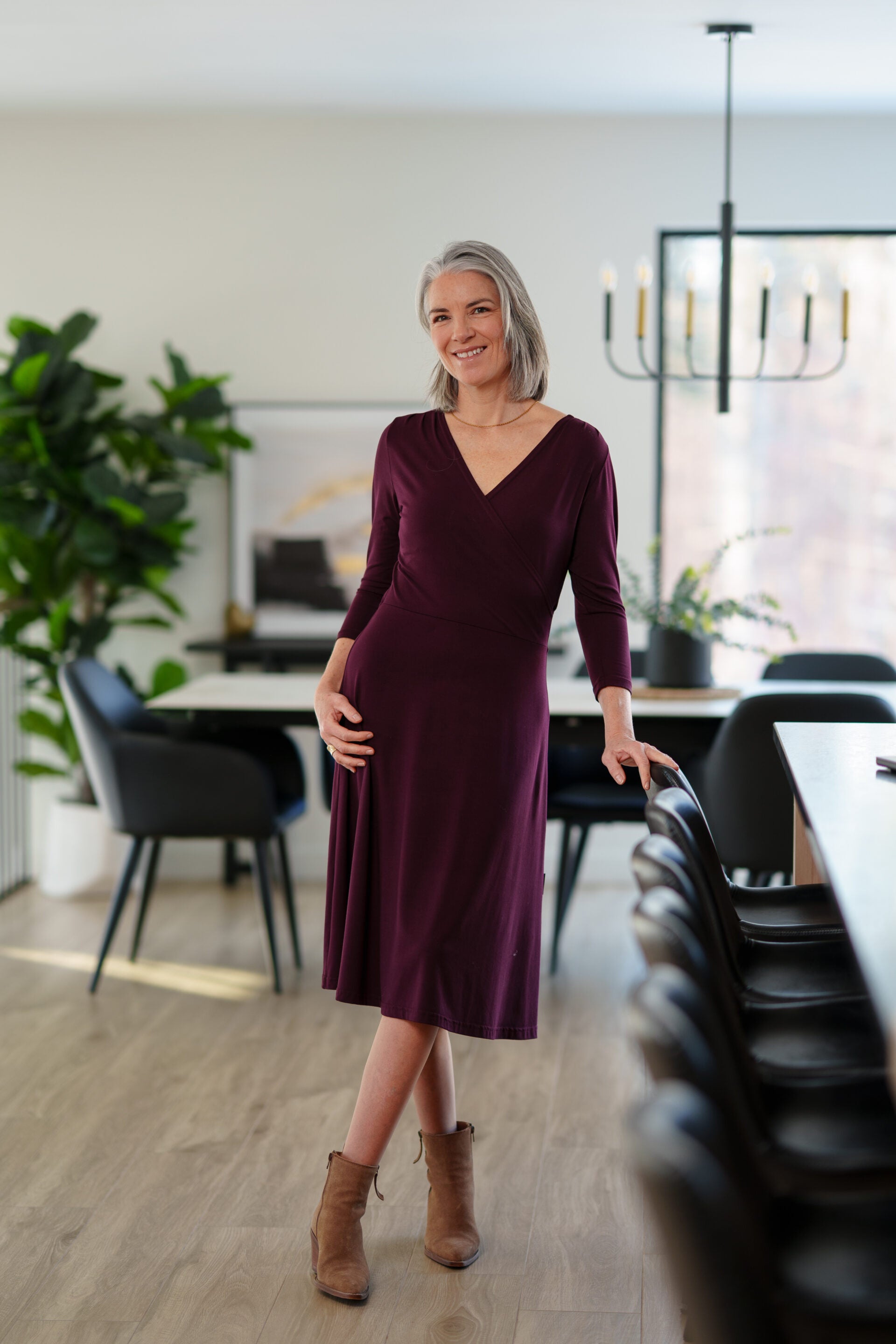 A woman wearing the St-Laurent Dress by Rien ne se Perd in Porto, a midi-length wrap style dress with a 3/4 sleeves and subtle shoulder pleats. She is standing in a dining room in front of a table. 