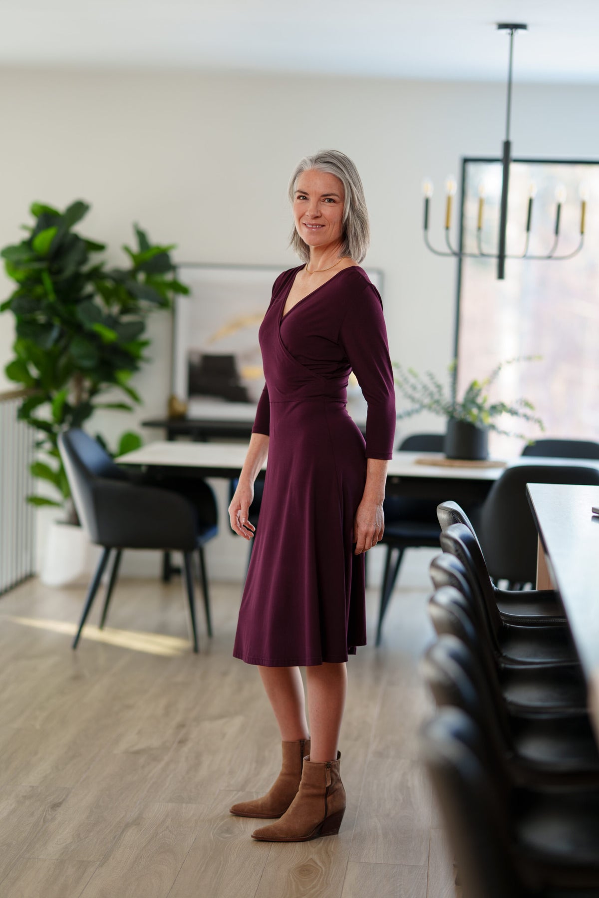 Side view of a woman wearing the St-Laurent Dress by Rien ne se Perd in Porto, a midi-length wrap style dress with a 3/4 sleeves and subtle shoulder pleats. She is standing in a dining room in front of a table. 