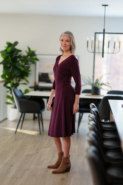 Side view of a woman wearing the St-Laurent Dress by Rien ne se Perd in Porto, a midi-length wrap style dress with a 3/4 sleeves and subtle shoulder pleats. She is standing in a dining room in front of a table. 