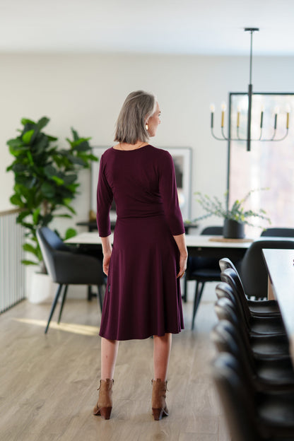 Back view of a woman wearing the St-Laurent Dress by Rien ne se Perd in Porto, a midi-length wrap style dress with a 3/4 sleeves and subtle shoulder pleats. She is standing in a dining room in front of a table. 