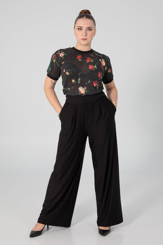 A woman wearing the Darling T-Shirt by Slak in Floral, a short sleeved, semi-fitted sweater with contrasting fabric at the cuffs, collar and hem, inserts on the sleeves, and a solid back. She is wearing it with black pants and standing in front of a white background. 