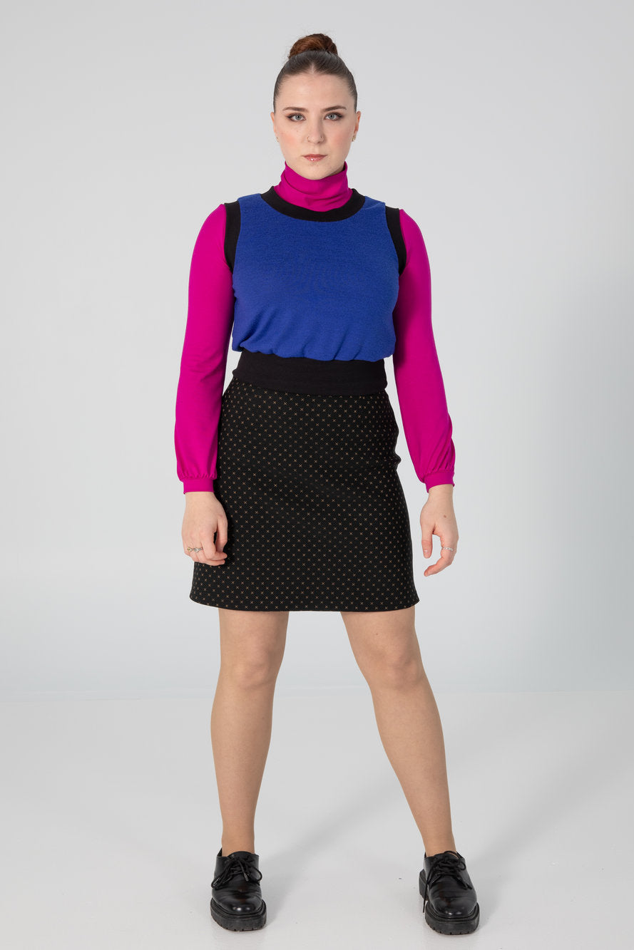 A woman wearing the Eve Skirt by Slak in Golden X print on Black, an above the knee-length pull-on skirt with a slight A-line shape. She is wearing it with a pink top and blue vest, and standing in front of a white background. 