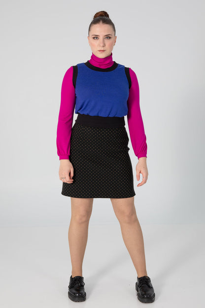 A woman wearing the Eve Skirt by Slak in Golden X print on Black, an above the knee-length pull-on skirt with a slight A-line shape. She is wearing it with a pink top and blue vest, and standing in front of a white background. 