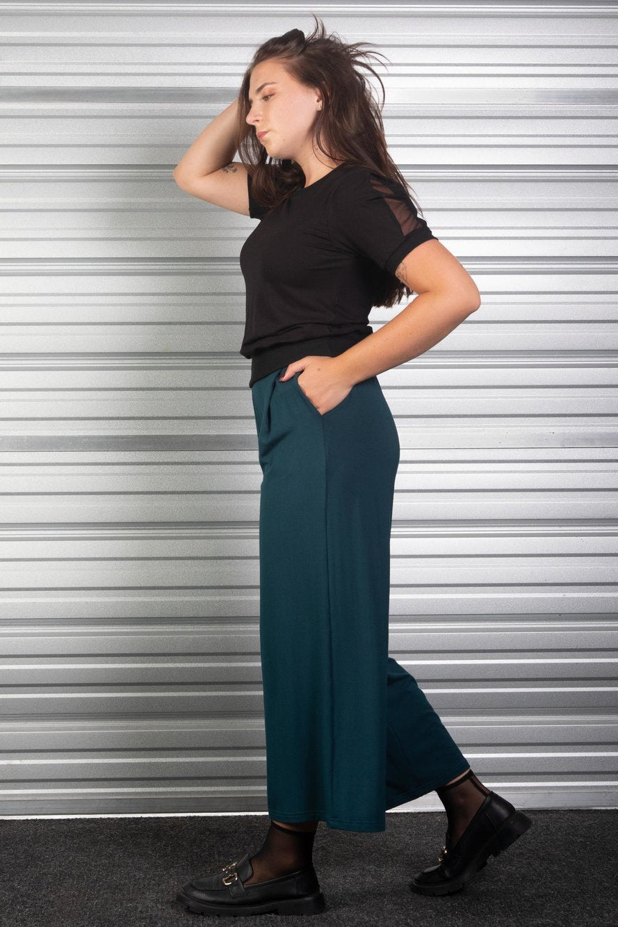 Side view of a woman wearing the Lotus Pants from Slak in Evergreen, wide-legged slightly cropped pants in viscose with an elastic waist, slanted pockets, and front and back pleats. She is wearing them with a colour blocked top of the and standing in front of a metallic shuttered door. 