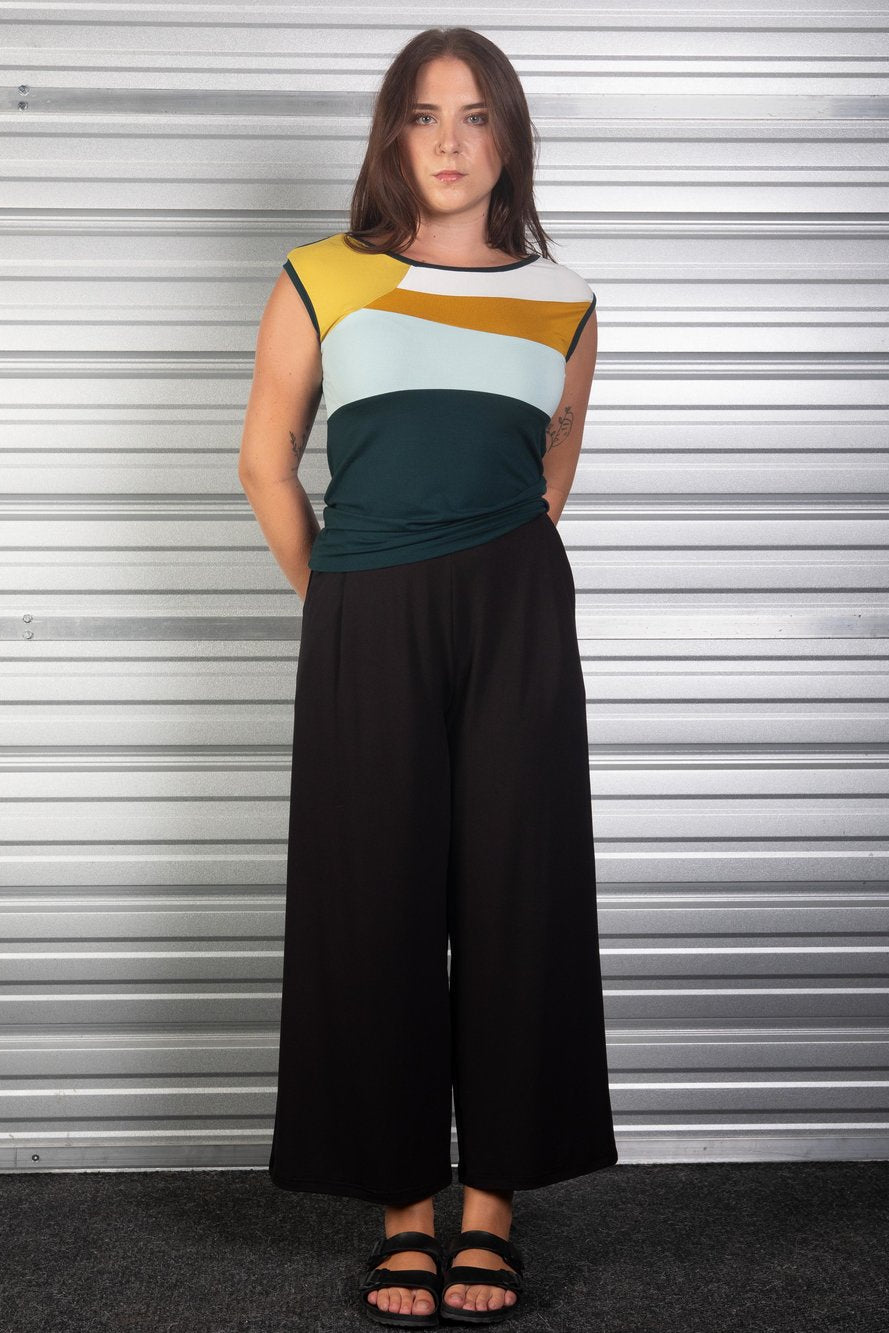 A woman wearing the Lotus Pants from Slak in Black, wide-legged pants in viscose with an elastic waist, slanted pockets, and front and back pleats. She is wearing them with a colour blocked top of the and standing in front of a metallic shuttered door. 