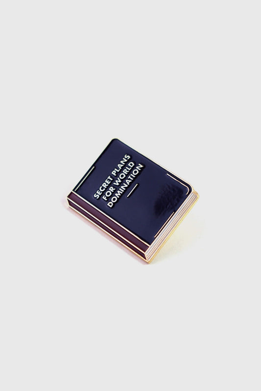 The World Domination Pin from Badaboöm Studio depicts a tiny book titled "Secret Plans for World Domination." It's shown against a white background. 