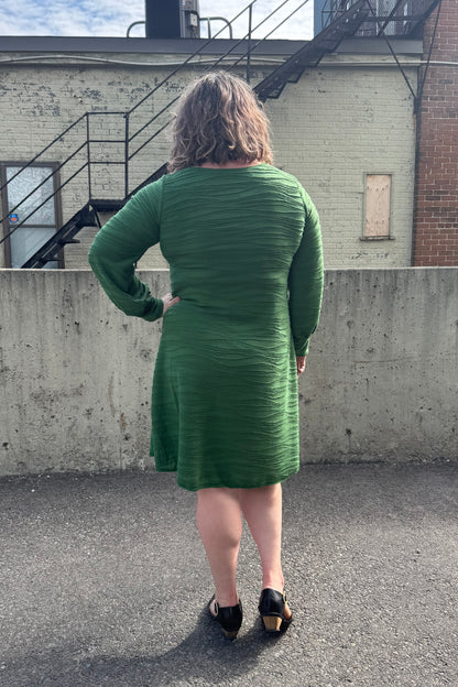 Back view of a woman wearing the Long Sleeved Casual Dress FW by SI Design in Green, with a round neck, long cuffed sleeves, and a loose A-line shape. she is standing in front of a building with a staircase.