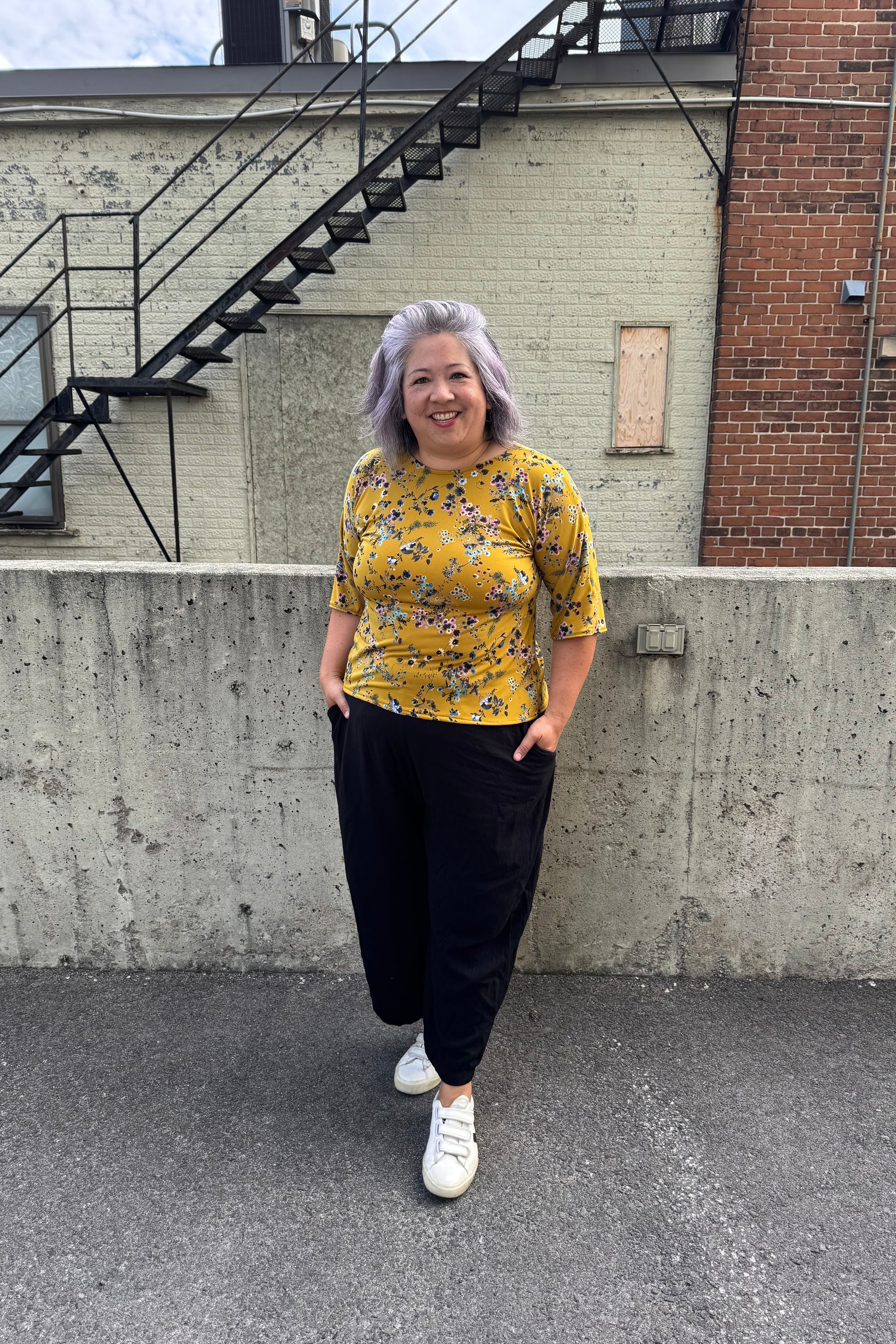 A woman wearing the Oversized Top by SI Design in Yellow Floral, featuring a scoop neck, elbow-length sleeves, and a slightly relaxed fit. She is wearing with black pants and standing outside in front of a brick building. 