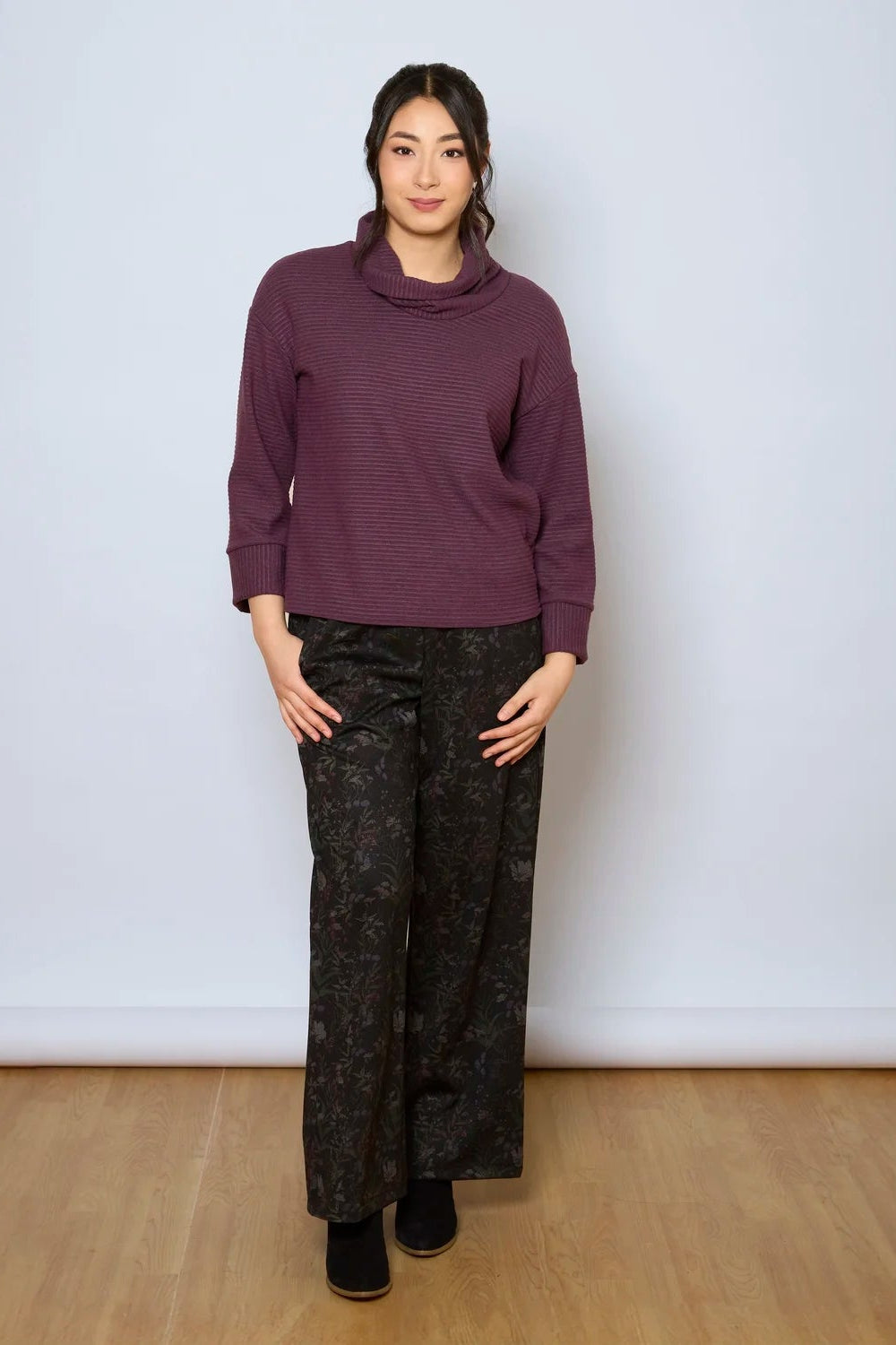 A woman wearing the Sidney Top by Tangente in Purple, a slightly cropped, boxy top in ribbed fabric with long cuffed sleeves and a large cowl neck. She is wearing it with floral pants and standing in front of a white wall. 