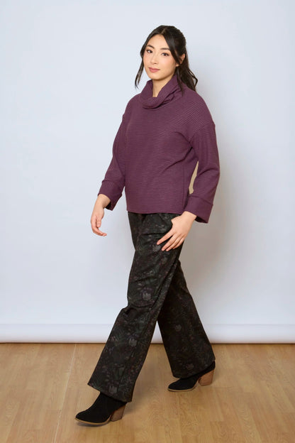 A woman wearing the Sidney Top by Tangente in Purple, a slightly cropped, boxy top in ribbed fabric with long cuffed sleeves and a large cowl neck. She is wearing it with floral pants and standing in front of a white wall. 