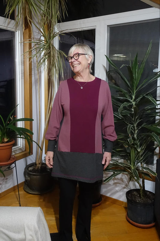 A woman wearing the Viola Tunic by Solomia in Burgundy/Black/Old Rose, a colour-blocked piece featuring long,  a round neckline, and a mid-thigh length. She is wearing it with black pants and standing in a room with plants.  