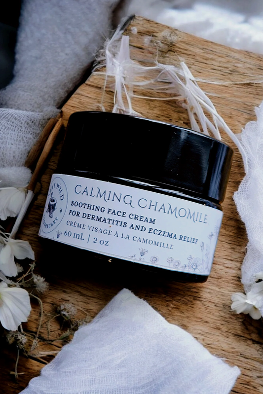 A jar of 'Calming chamomile' face cream by Stay Wild Apothecary is shown on a wooden board with white flowers