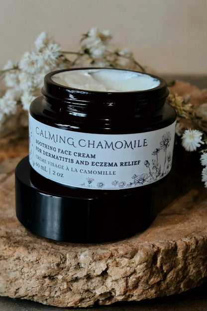 A jar of 'Calming chamomile' face cream by Stay Wild Apothecary is shown on a wooden board with white flowers