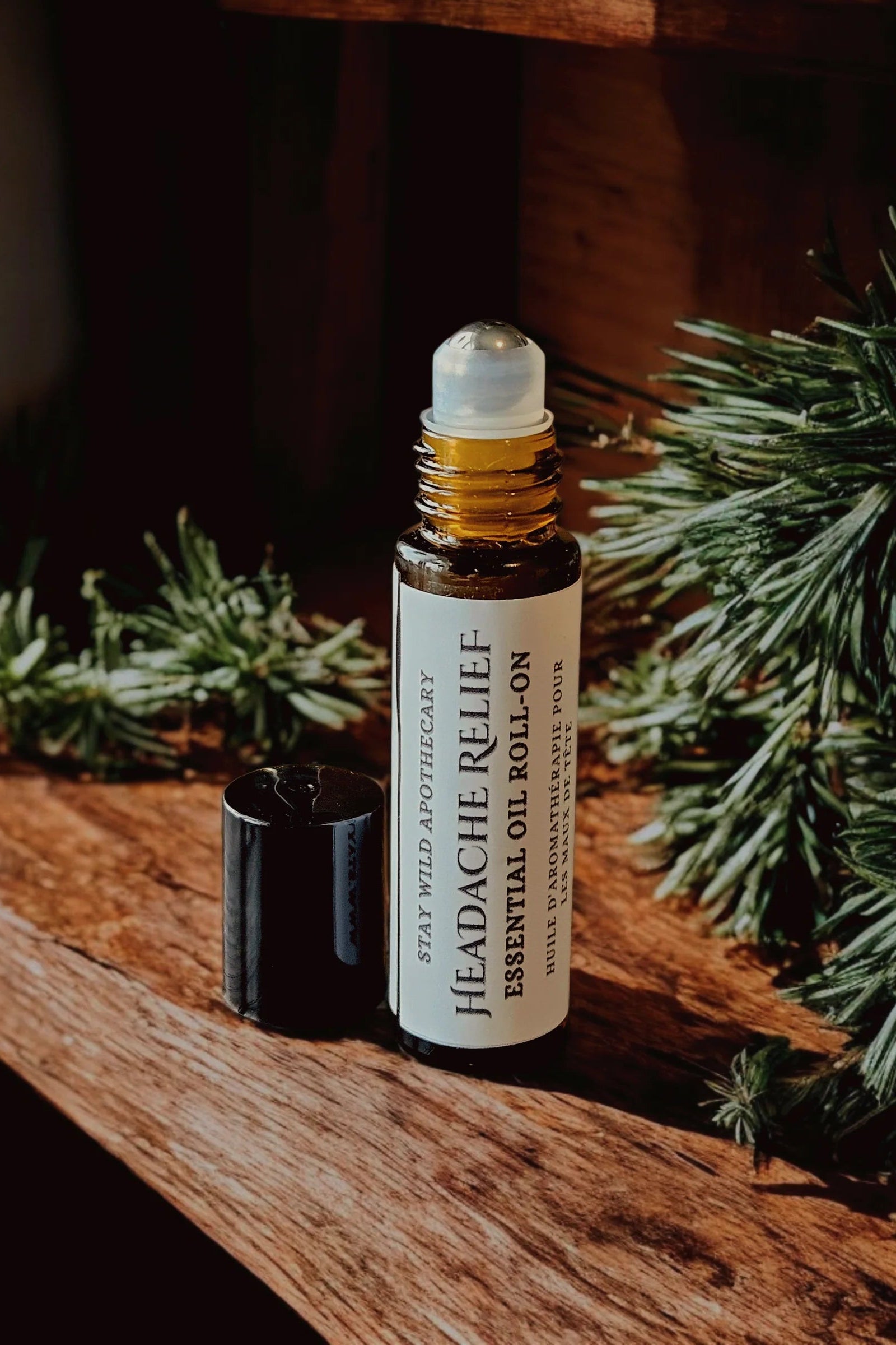 A bottle of 'Headache Relief'  essential oil roll-on by Stay Wild Apothecary is shown on a wooden surface with greenery.