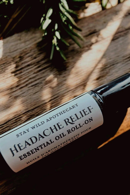 A bottle of 'Headache Relief'  essential oil roll-on by Stay Wild Apothecary is shown on a wooden surface with greenery.