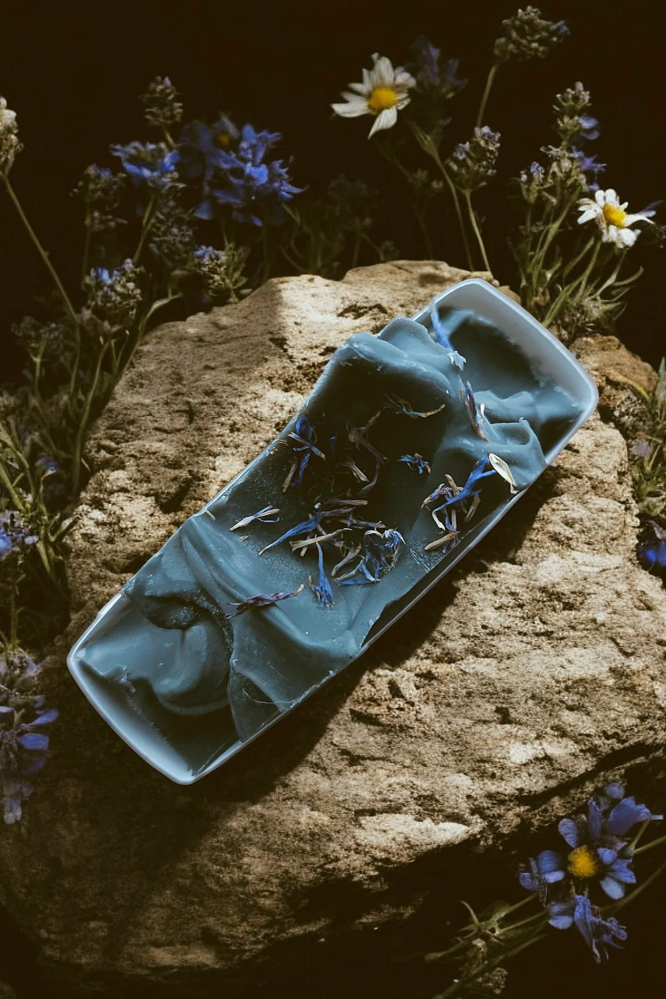 A bar of 'Spellbound' soap by Stay Wild Apothecary is shown on a stone with flowers surrounding it. 