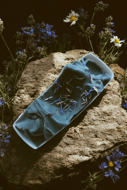 A bar of 'Spellbound' soap by Stay Wild Apothecary is shown on a stone with flowers surrounding it. 