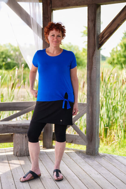 A woman wearing the Modern T-Shirt by Rien ne se Perd in Blue, a bamboo tee with a round neck, short-sleeves, and a tie at the waist. She is wearing them with a skirt and leggings and is standing outside in a wooden shelter. 