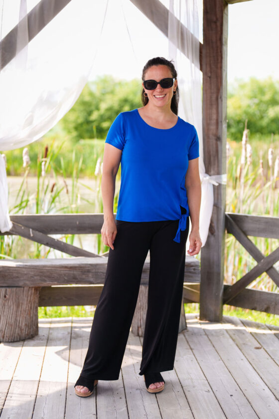 A woman wearing the Modern T-Shirt by Rien ne se Perd in Blue, a bamboo tee with a round neck, short-sleeves, and a tie at the waist. She is wearing them black pants and is standing outside in a wooden shelter. 