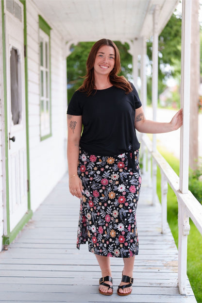 A woman wearing the Fiesta 7/8 Skirt by Rien ne se Perd in Colourful Daisies, a slightly A-line skirt with an elastic waist and side slits. She is wearing it with a black top and standing on a wooden porch.