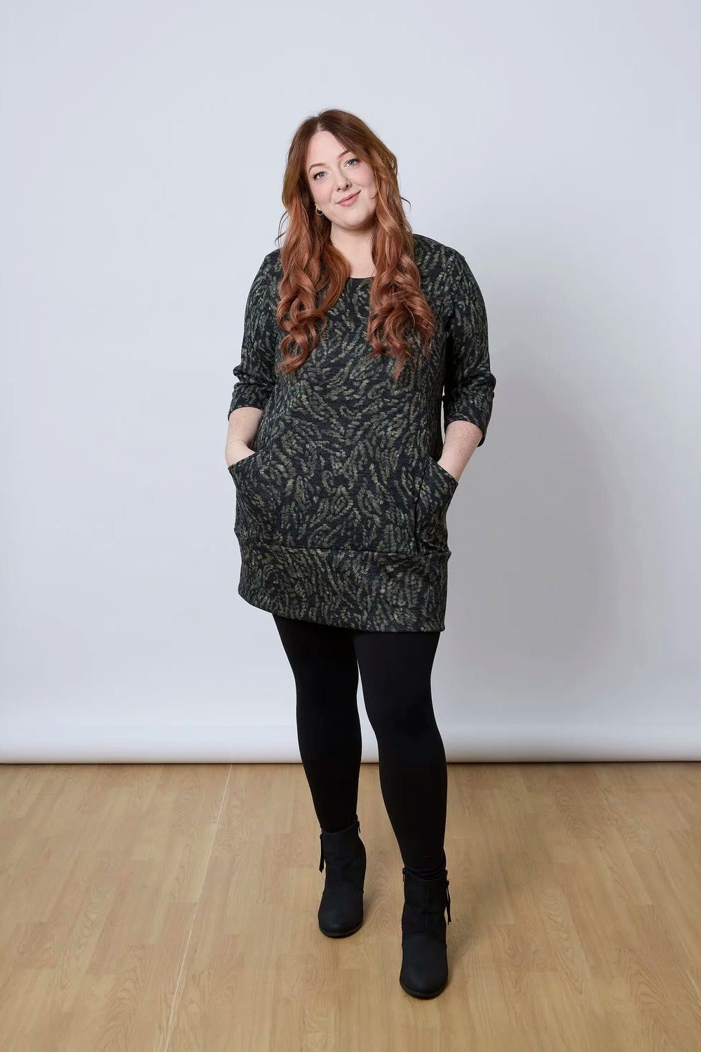 A woman wearing the Brooke Tunic by Tangente in Green Print, with 3/4 sleeves and a fitted band at the bottom. She is wearing it with black leggings and standing against a white wall. 