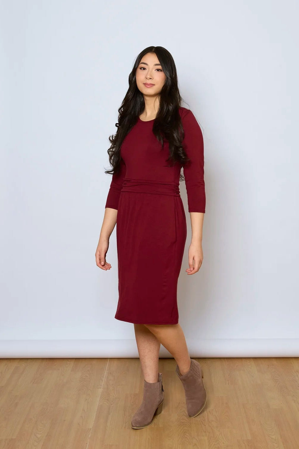 A woman wearing the Cascade Dress by Tangente in Burgundy, with a round neck, 3/4 sleeves, gathered defining waistband, and a gathered knee-length skirt. She is standing against a white wall.