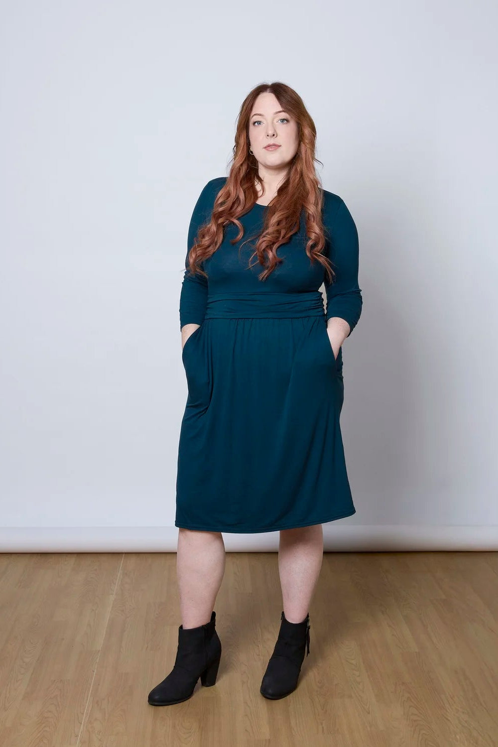 A woman wearing the Cascade Dress by Tangente in Peacock, with a round neck, 3/4 sleeves, gathered defining waistband, and a gathered knee-length skirt. She is standing against a white wall.