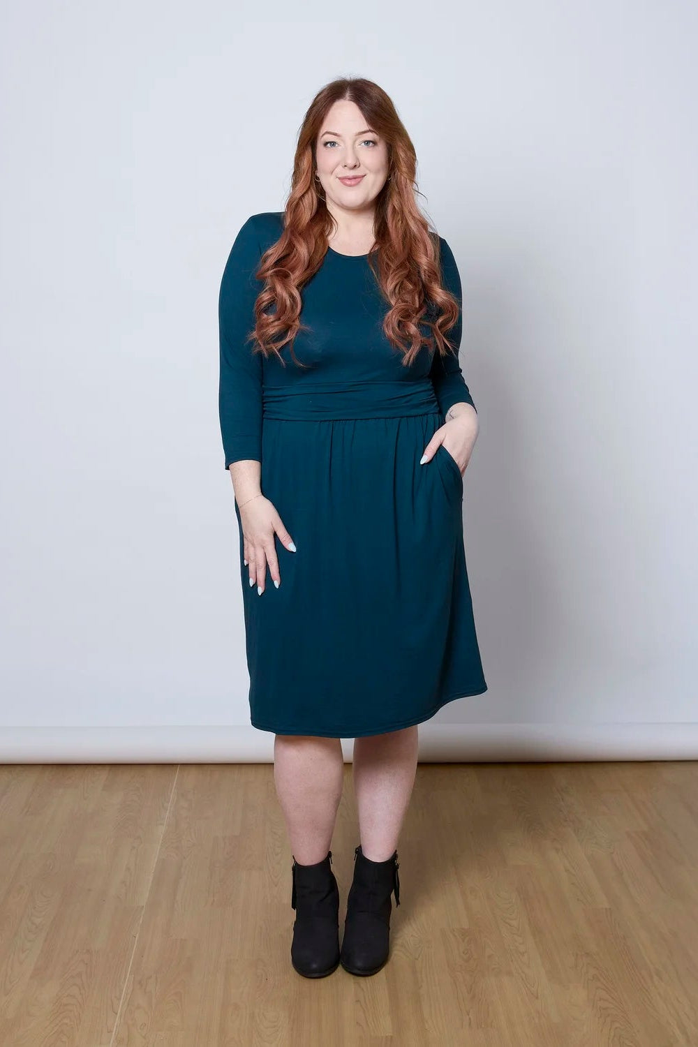 A woman wearing the Cascade Dress by Tangente in Peacock, with a round neck, 3/4 sleeves, gathered defining waistband, and a gathered knee-length skirt. She is standing against a white wall.