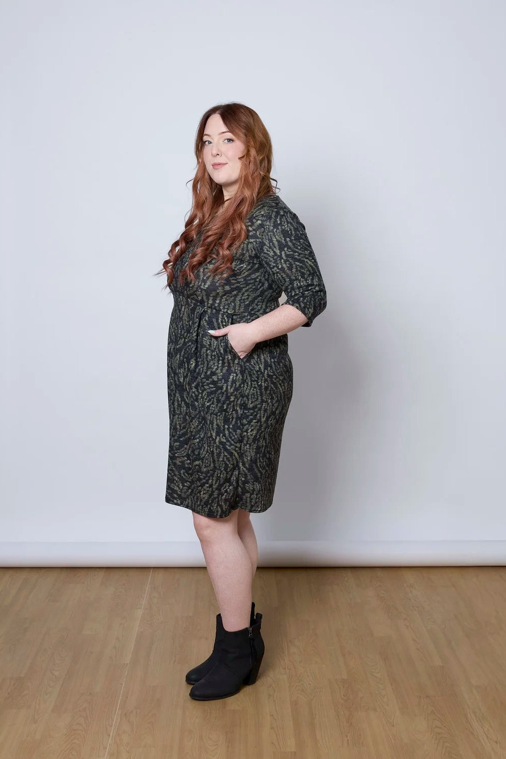 Side view of a woman wearing the Meadow Dress by Tangente in Green Print, with a round neck, 3/4 sleeves, princess seams, and a pleated knee-length skirt. She is standing against a white wall. 