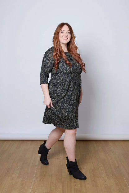 A woman wearing the Meadow Dress by Tangente in Green Print, with a round neck, 3/4 sleeves, princess seams, and a pleated knee-length skirt. She is standing against a white wall. 