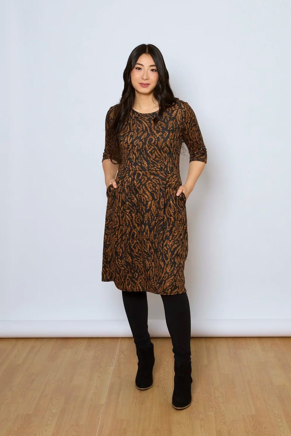 A woman wearing the Meadow Dress by Tangente in Rust Print, with a round neck, 3/4 sleeves, princess seams, and a pleated knee-length skirt. She is standing against a white wall. 