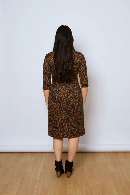 Back view of a woman wearing the Meadow Dress by Tangente in Rust Print, with a round neck, 3/4 sleeves, princess seams, and a pleated knee-length skirt. She is standing against a white wall. 
