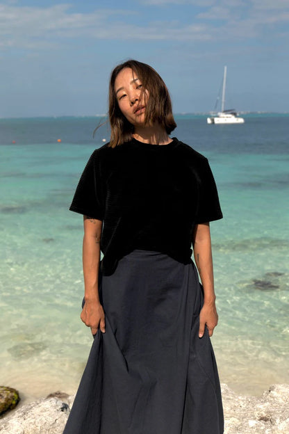 A woman wearing the Tofino T-Shirt by MAS in Pepper, a short sleeved top in ribbed velvet. She is wearing it with a blue skirt and standing on the beach. 