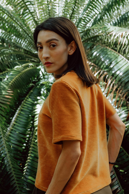 A woman wearing the Tofino T-Shirt in Tumeric by MAS, a short sleeved top in ribbed velvet. She is standing in front of green foliage.