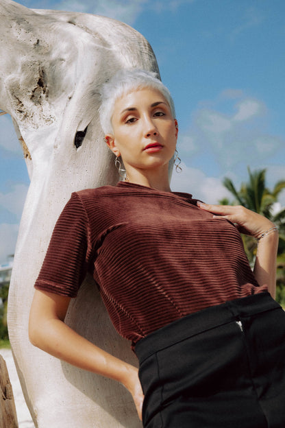 A woman wearing the Tofino T-Shirt by MAS in Cacao, a short sleeved top in ribbed velvet. She is standing on a beach.