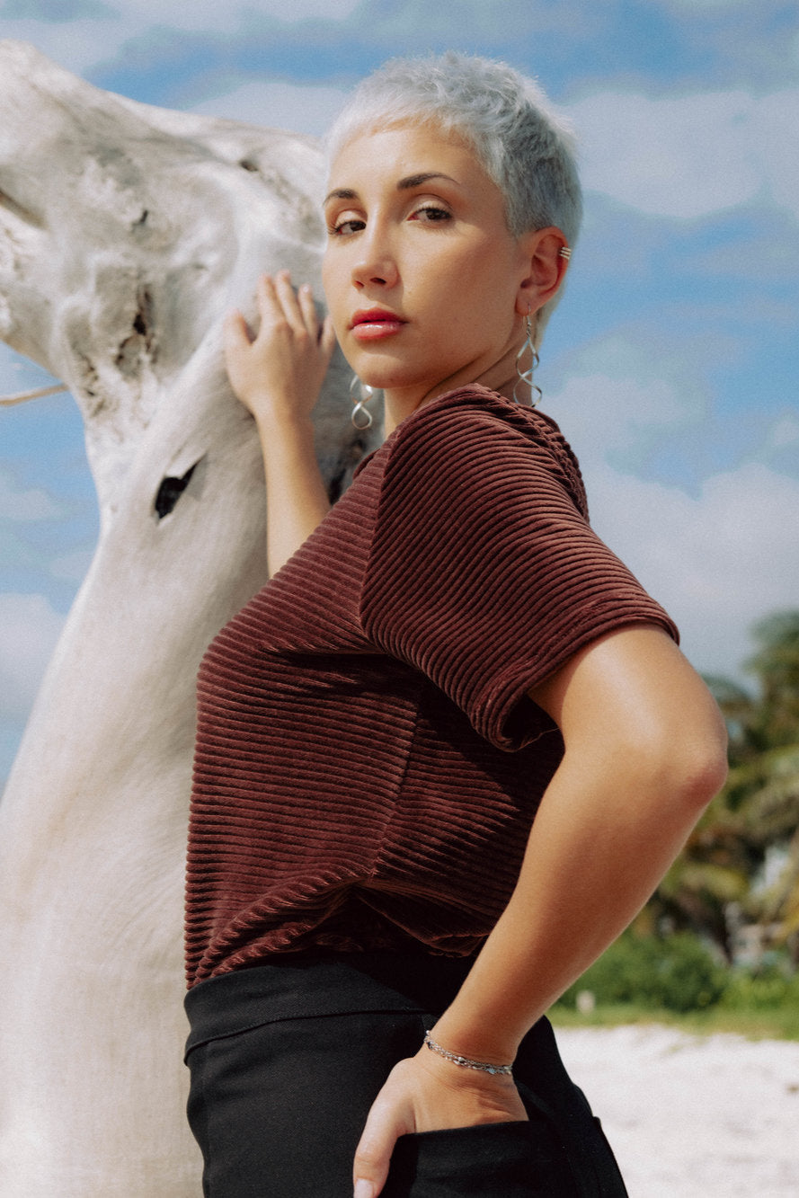 Side view of a woman wearing the Tofino T-Shirt by MAS in Cacao, a short sleeved top in ribbed velvet. She is standing on a beach.