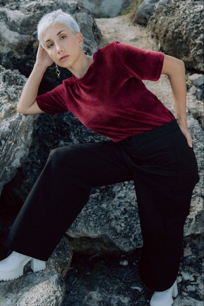 Back view of a woman wearing the Tofino T-Shirt by MAS in Rosewood, short sleeved top in ribbed velvet. She is wearing it with black pants and standing on rocks.
