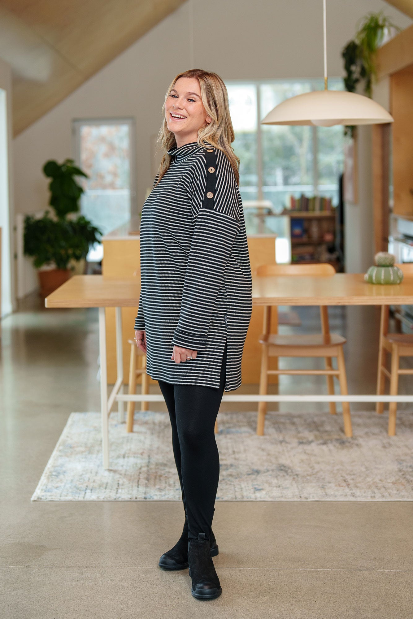 Side view of a woman wearing the Aralie Tunic by Rien ne se Perd in Black and White Stripe, with a high neckline, long sleeves, side slits for ease, drop sleeves and a coconut button placket on the left shoulder. She is wearing it with black leggings and standing in a kitchen. 