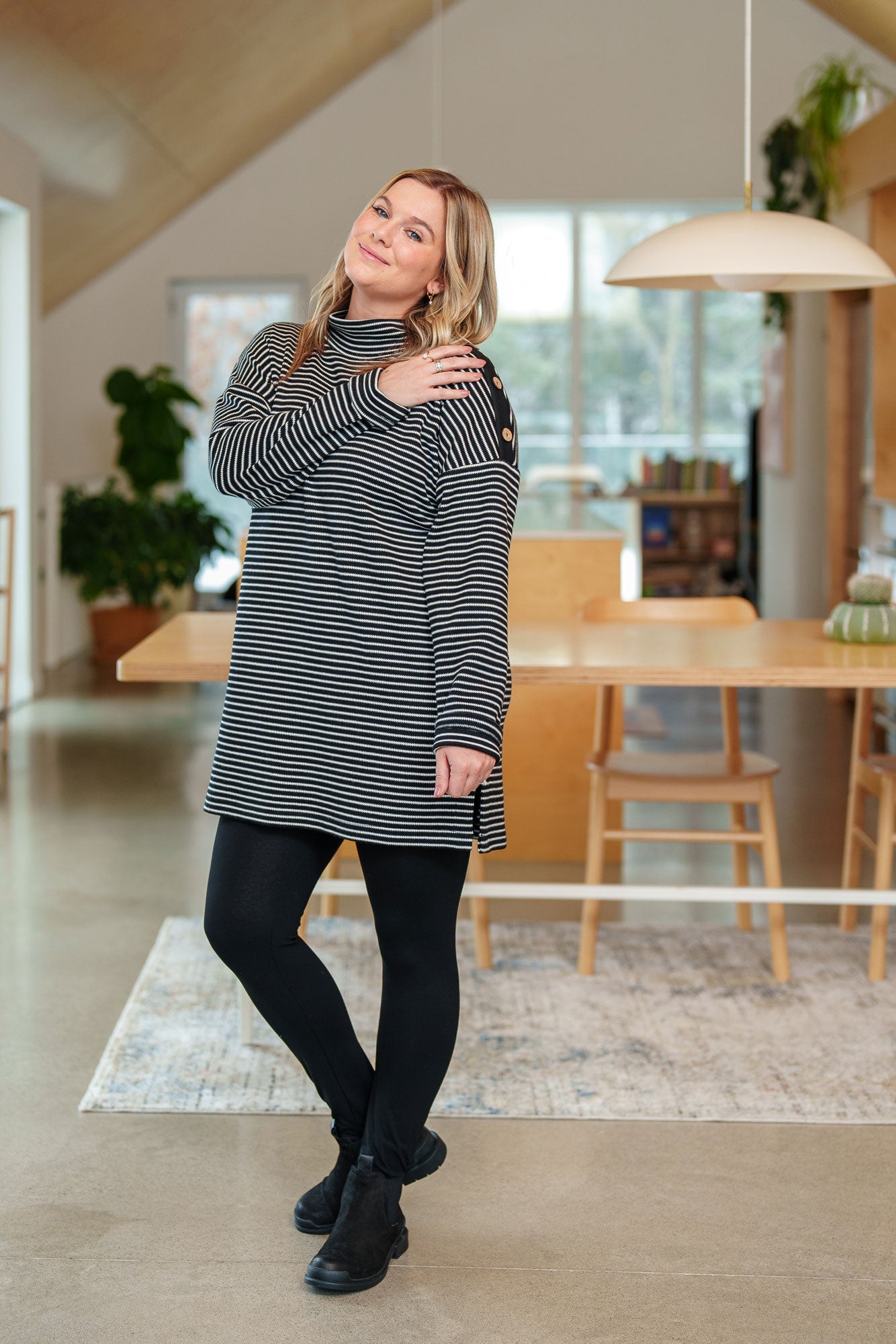 A woman wearing the Aralie Tunic by Rien ne se Perd in Black and White Stripe, with a high neckline, long sleeves, side slits for ease, drop sleeves and a coconut button placket on the left shoulder. She is wearing it with black leggings and standing in a kitchen. 