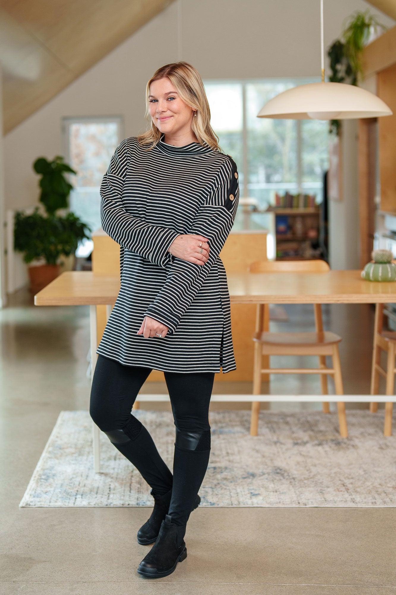 A woman wearing the Aralie Tunic by Rien ne se Perd in Black and White Stripe, with a high neckline, long sleeves, side slits for ease, drop sleeves and a coconut button placket on the left shoulder. She is wearing it with black leggings and standing in a kitchen. 