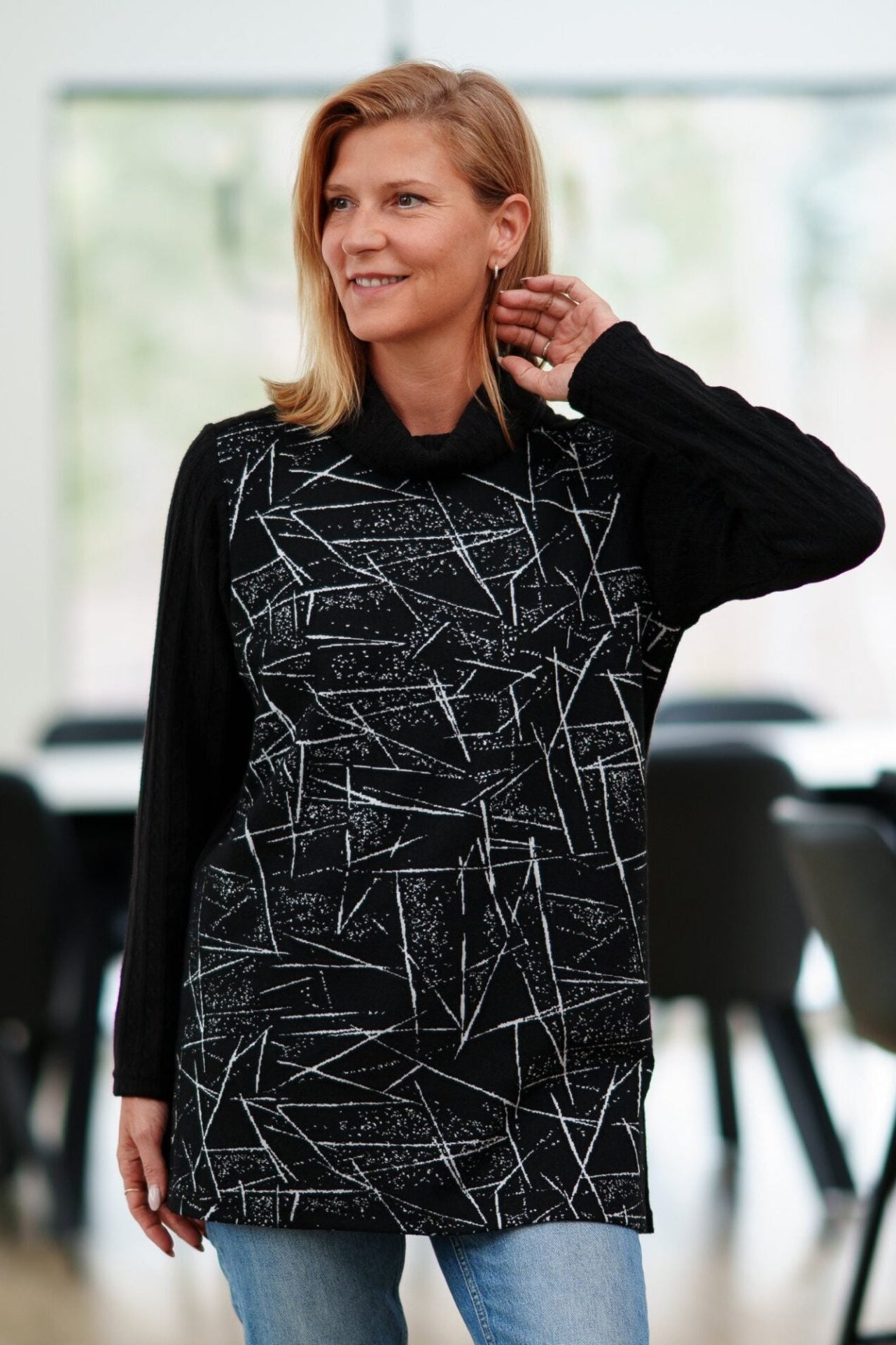 A woman wearing the Auclair Tunic by Rien ne se Perd in a black and white geometric print, with a solid black turtleneck, back, and sleeves, and side slits. She is wearing it with jeans and standing in a room with indoors with a dining room table behind her. 