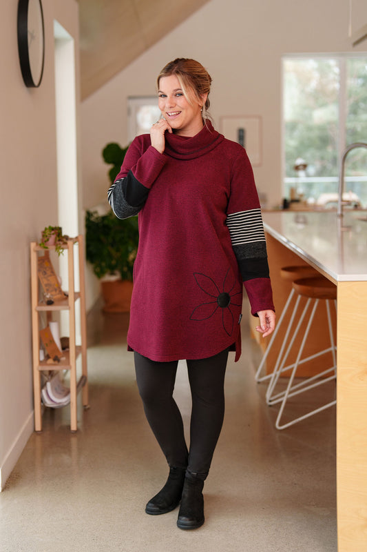 A woman wearing the Églantier Tunic by Rien ne se Perd in textured Bordeaux fabric with striped and black colour-blocking on the long sleeves. It has a large turtleneck, a faux leather floral decal, and a side pocket. She is wearing it with black leggings and standing in a kitchen. 