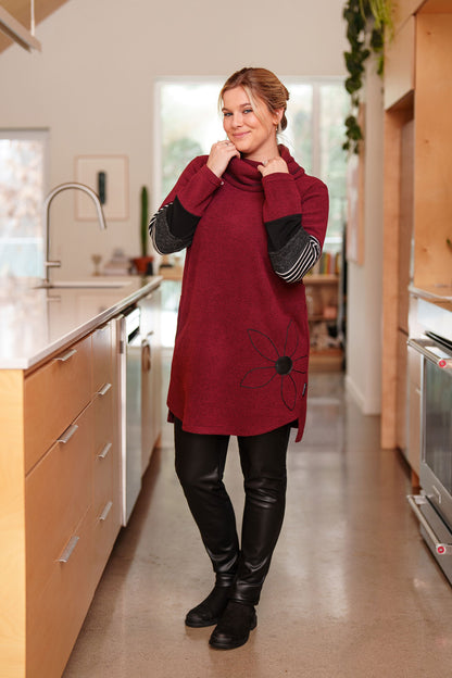 A woman wearing the Églantier Tunic by Rien ne se Perd in textured Bordeaux fabric with striped and black colour-blocking on the long sleeves. It has a large turtleneck, a faux leather floral decal, and a side pocket. She is wearing it with black leggings and standing in a kitchen. 