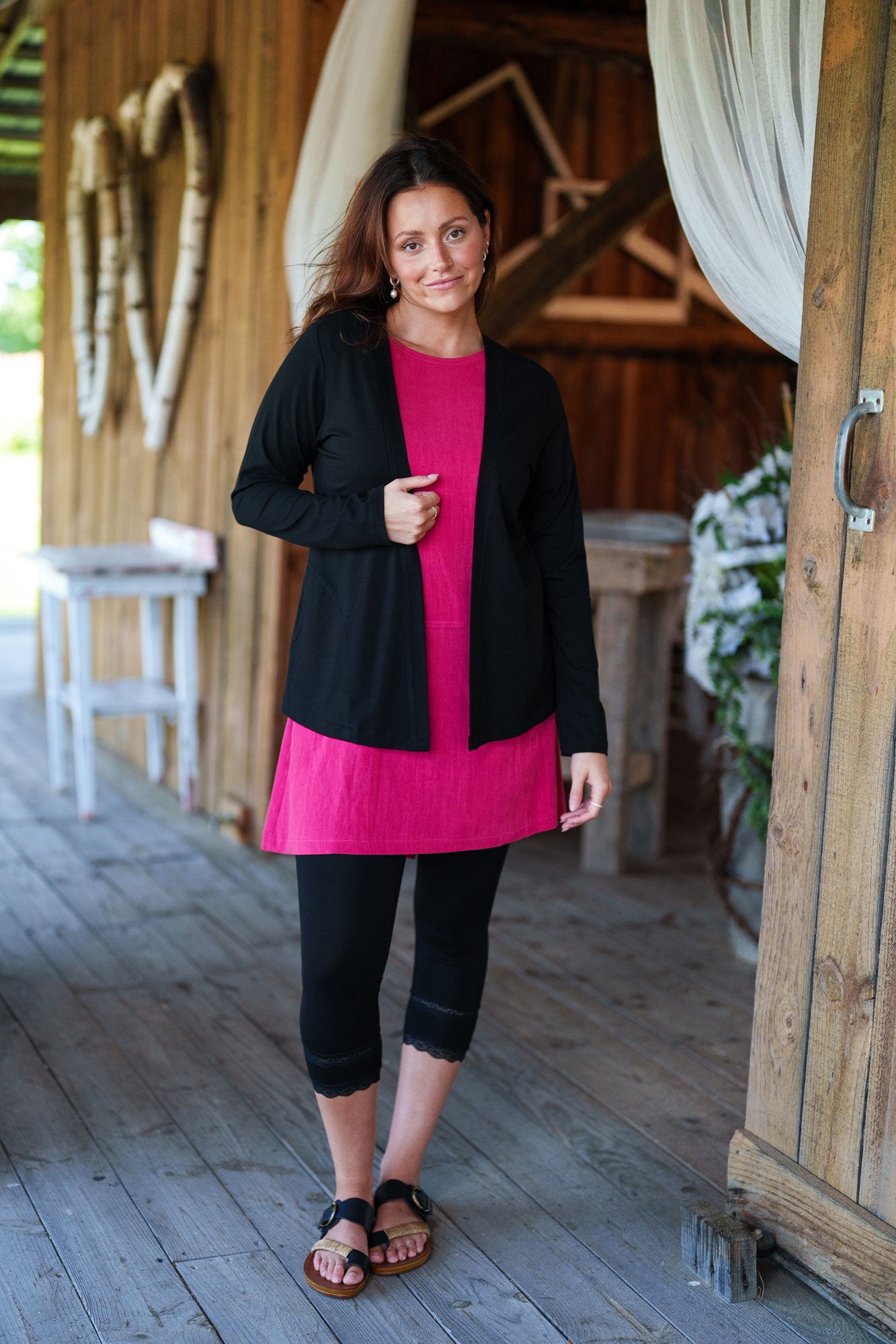 3/4 shot of a woman wearing the Merengue Tunic by Rien ne se Perd in Raspberry, a loose-fitting linen and viscose tunic with a round neck, short sleeves, asymmetrical cut, and patch pocket. She is wearing it with black capri leggings and a black bolero and is standing outside.