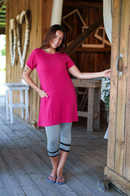 3/4 shot of a woman wearing the Merengue Tunic by Rien ne se Perd in Raspberry, a loose-fitting linen and viscose tunic with a round neck, short sleeves, asymmetrical cut, and patch pocket. She is wearing it with black and white leggings and is standing outside.