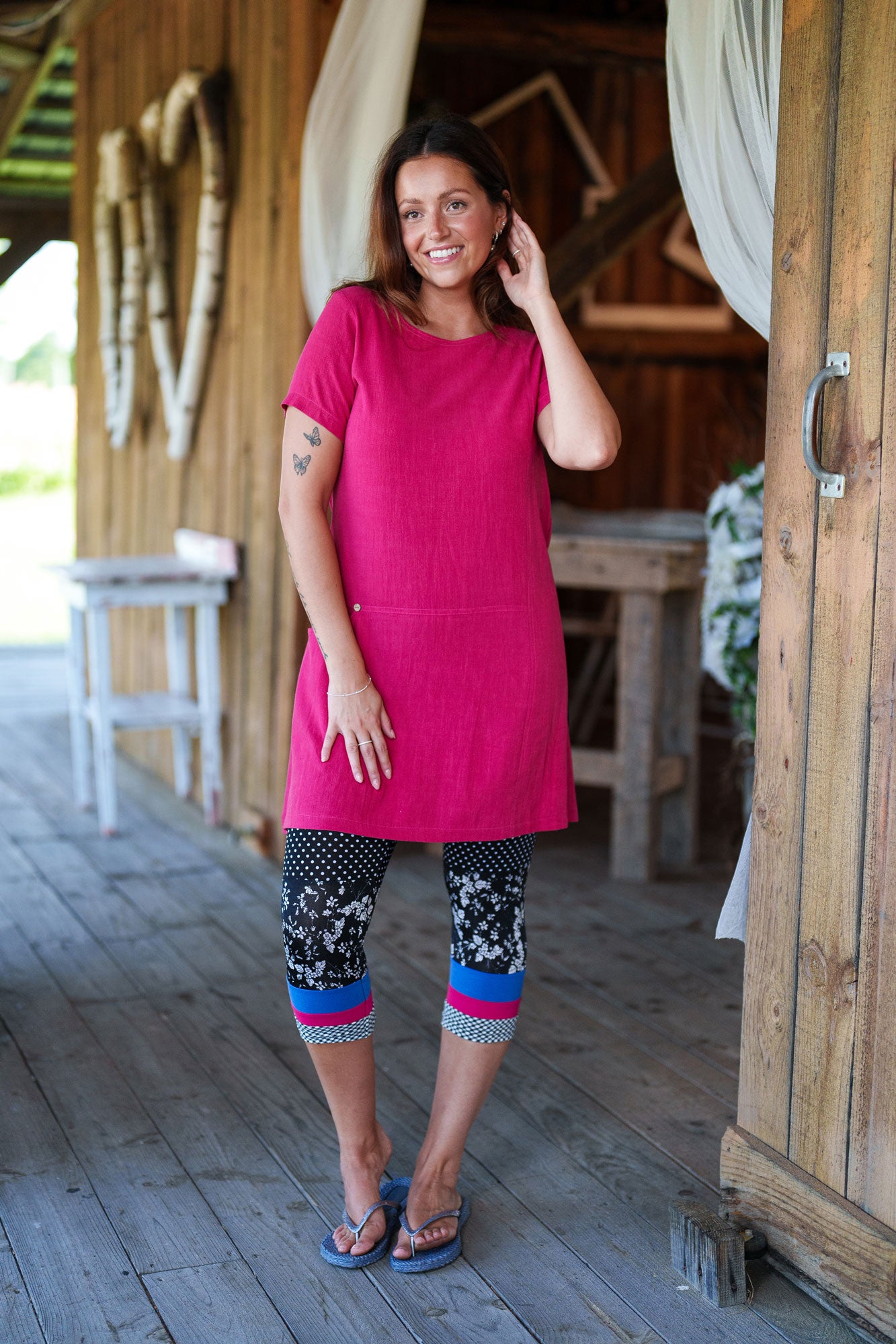 3/4 shot of a woman wearing the Merengue Tunic by Rien ne se Perd in Raspberry, a loose-fitting linen and viscose tunic with a round neck, short sleeves, asymmetrical cut, and patch pocket. She is wearing it with patterned leggings and is standing outside on a porch.